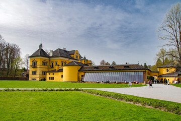 Obraz premium Hellbrunn Palace in Salzburg with bright yellow facade, green park lawn and visitors under dramatic cloudy sky in Austria