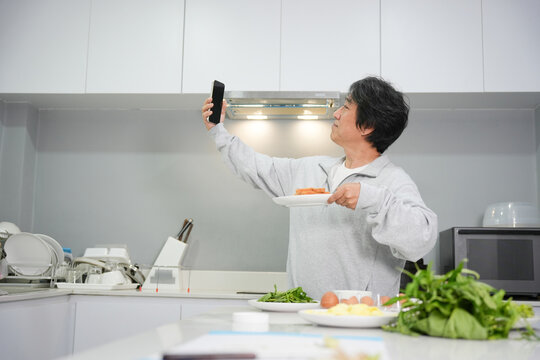 Mature Man Taking a Selfie with His Homemade Meal, Asian Man Documenting His Cooking or Food Preparation on a Smartphone, Middle-Aged Man Posing with a Plate of Food in a Kitchen