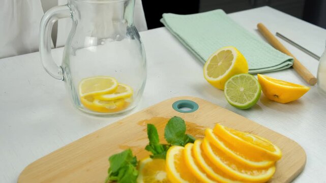 Woman hands preparing lemonade putting citrus slices into glass jug on kitchen at home. Preparing fresh homemade vitamin drink with organic products, herbs mint, melissa. Recipe cuisine culinary.