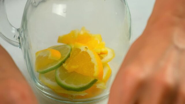 Preparing lemonade putting citrus slices into glass jug on kitchen at home. Preparing fresh homemade vitamin drink with organic products, herbs mint, melissa. Recipe cuisine culinary.