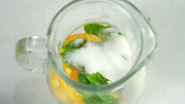 Preparing lemonade putting sugar to herb mint, citrus slices into glass jug. Cooking fresh homemade vitamin drink with organic products on kitchen at home. Recipe cuisine culinary ingredient.