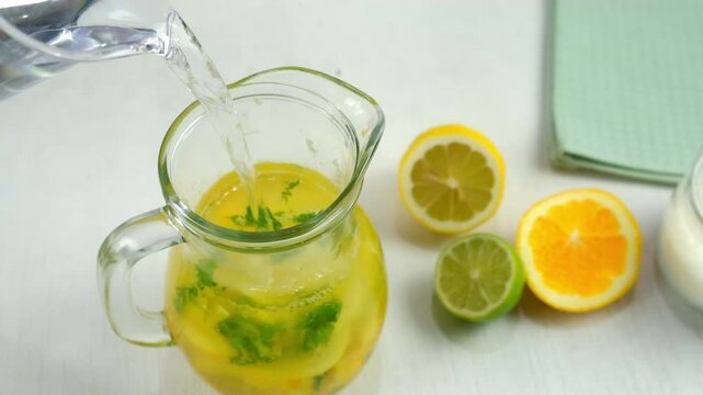 Preparing homemade lemonade pouring water to citrus fruit herbs in glass jug. Cooking mixing herb mint, orange, lime, lemon with sugar on kitchen at home. Recipe cuisine culinary ingredient.