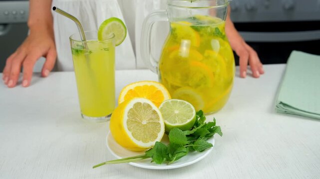 Woman with homemade healthy lemonade from citrus fruit mint herbs on table. Cooking fresh organic beverage with herb mint orange lime lemon on kitchen at home. Recipe cuisine culinary ingredient.