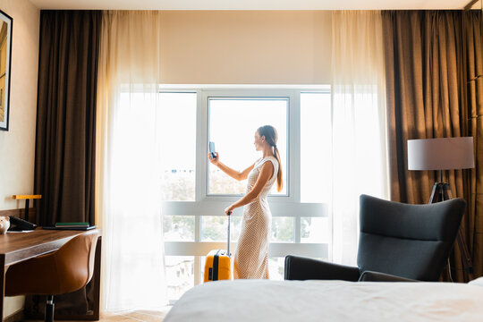 Woman with suitcase standing by hotel window taking picture of city view.