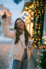 Fototapeta premium A girl in winter clothes walks through the evening New Year's city, drinking coffee on the street