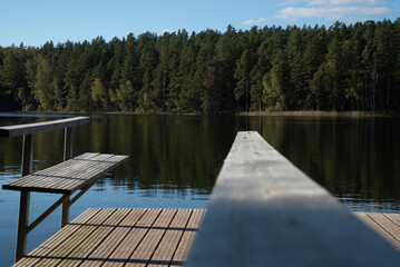 Naklejka premium Wooden pier with benches extends over a calm forest lake, with dense green trees reflecting in the still water under a clear blue sky.
