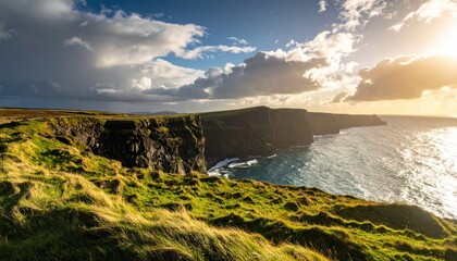 Dramatic Cliffs Overlooking the Ocean with a Cloudy Sky at Sunrise Sunlight Hitting the Water with Green Grass in the Foreground and Dark Rocks