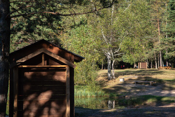 A small wooden shed in a forest clearing with trees, picnic benches, and a fire pit in the...
