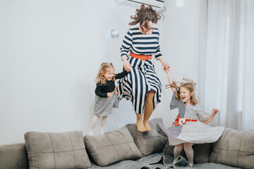 Mother and daughters happily jumping on their living room couch, sharing joyful family moments of bonding and laughter.