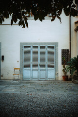 A charming, rustic scene in a quiet Florentine alley, featuring a weathered blue shuttered door, an empty wooden chair, and ancient stone textures bathed in soft, evocative light.