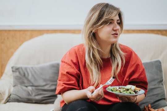 A young woman in a red shirt enjoying a plate of fresh salad while conversing on a couch. The casual indoor setting conveys a relaxed and friendly atmosphere.