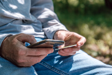 young adult sits on wooden bench in park, holding smartphone in one hand and card in other. sunlight filters through leaves above, creating serene atmosphere. close up.