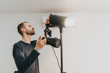 A man preparing photography equipment in a well-lit studio, ensuring proper lighting for a shoot. He holds a camera while adjusting the position and settings of a studio light stand.