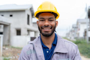 Portrait of a smiling construction engineer wearing a yellow hard hat and safety uniform at a housing development site. Confident male worker stands outdoors at a modern building construction area.