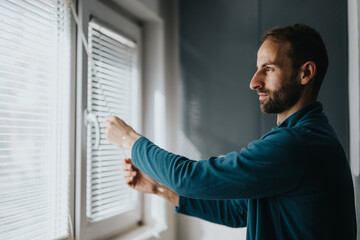 A man is seen adjusting horizontal window blinds in his bright and stylish apartment. This activity demonstrates an aspect of everyday life and home decor.
