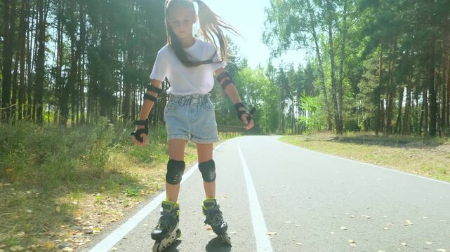 Child girl in casual clothes roller skating on road in forest on sunny summer day.