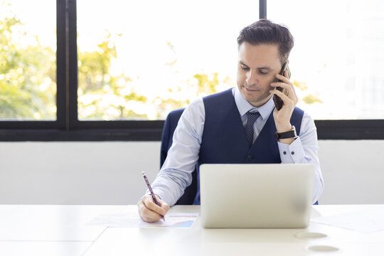 Businessman multitasking with smartphone call, writing notes, and working on laptop at office desk, representing communication, productivity, remote work, strategy planning, and corporate lifestyle.
