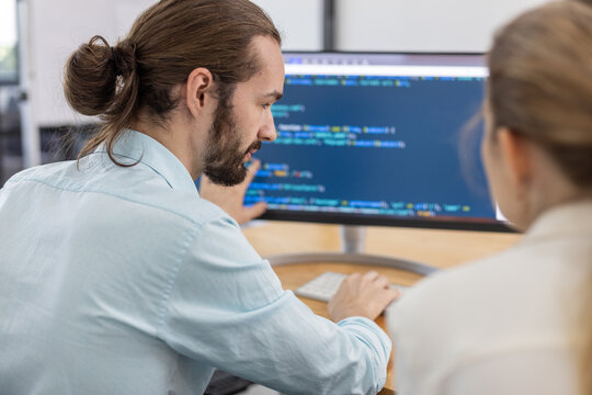 Software developer explaining code on computer screen to colleague in modern office, representing teamwork, programming, collaboration, software development, and tech industry environment.