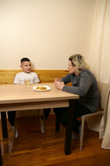 Family moment with mother and son sitting at table, drinking tea, eating cookies, and enjoying warm conversation together indoors.
