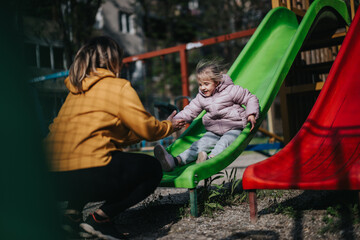 A mother assists her daughter sliding down a green slide at a playground, fostering joy.
