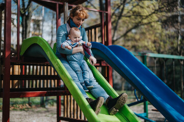 Obraz premium A woman and her child share a joyful moment sliding down a playground structure. Bonding, smiles, and laughter are captured in this vibrant outdoor scene under the sun.