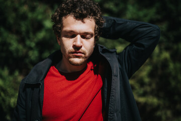 A man with a thoughtful expression outdoors, wearing a red shirt and black jacket, surrounded by lush greenery.