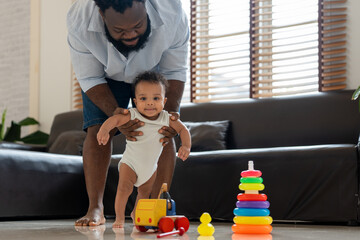 Happy African American dad holding adorable infant son helps him take first steps walking in living room at home.