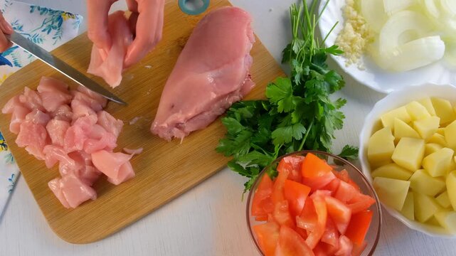 Chef woman hands cutting chicken breast into pieces on wooden board on kitchen. Bowl with ingredients near on table parsley, tomato, potato, garlic, onion. Cuisine, culinary, preparing domestic food.