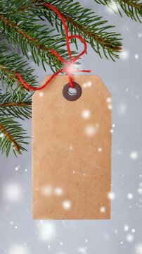 Blank kraft paper tag hanging on Christmas tree branch with red string, subtle camera movement and falling snow, festive winter background with copy space for holiday greetings and seasonal design pro