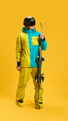 Skier holding skis upright in bright yellow studio with protective helmet and goggles. Concept of...