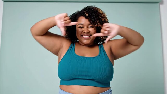 Energetic curvy woman in a teal sports bra smiles broadly against a pale backdrop, arms raised playfully over her eyes, curly hair framing her joyful, confident expression, embracing selfcare