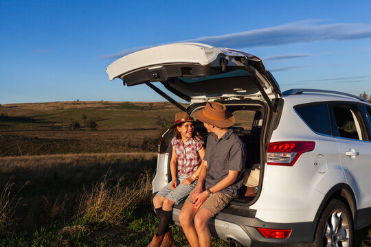 Teen and preteen Australian siblings sitting in car boot at rest stop in rural Australia
