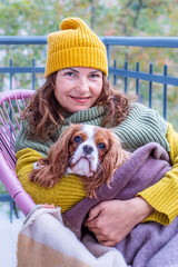 Midlife woman wearing a yellow beanie and cozy sweater sits on a balcony with her dog wrapped in a blanket, enjoying the autumn ambiance and companionship during menopause