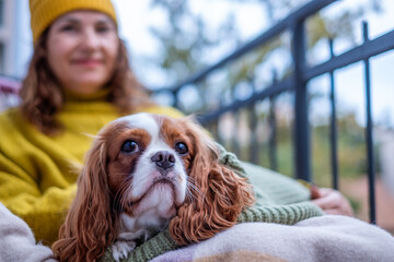 Midlife woman in cozy sweater and hat sits on balcony with dog, wrapped in blanket, enjoying autumn...