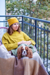 Woman in midlife wearing a cozy yellow sweater and hat sits on a balcony with her dog, wrapped in a blanket, enjoying the autumn ambiance and companionship during menopause