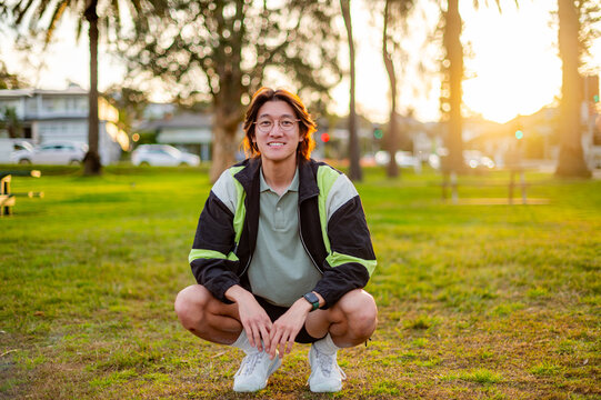 A male in athletic wear squats on grass, smiling as the sun sets behind the trees