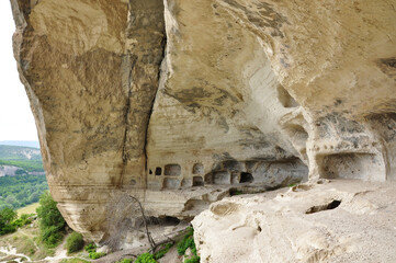 A huge natural rock overhang that reveals the remains of multi-tiered cave city architecture with numerous square and rectangular niches, set against a green valley.