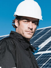 A man in work attire and a safety helmet stands sideways near a solar panel generating electricity. Production of clean energy.
