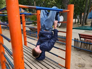 A boy dressed in a blue puffer jacket and jeans hangs upside down from the orange metal bars of a playground climbing frame, having fun in the park © Татьяна Швец