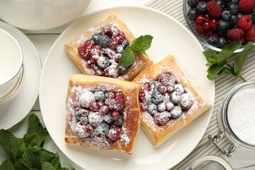 Tasty puff pastries with berries, powdered sugar and mint on white wooden table, flat lay