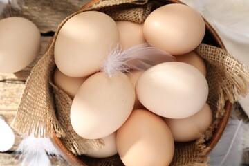 Raw chicken eggs in bowl and feathers on wooden table, top view