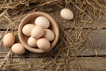 Raw chicken eggs in bowl and straw on wooden table, flat lay. Space for text