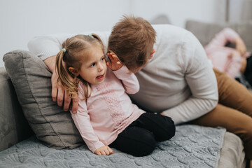 A kind father shares a tender moment with his daughter while sitting on the couch at home. Their interaction highlights family connection, love, and care during a moment of relaxation.