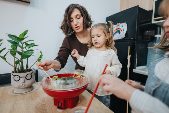 A mother assists her young daughters while making homemade cotton candy in the kitchen. They are enjoying a fun and creative bonding time together. The scene is warm and filled with joy.
