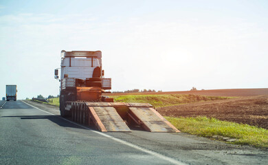 An empty lowboy truck stands on the road for loading equipment against the background of a field, industry. Copy space for text