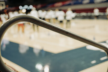 Curved steel handrail in sharp focus leads into a basketball college stadium in Dallas, Texas. Blurred figures in white shirts move across hardwood, blending infrastructure with athletic energy
