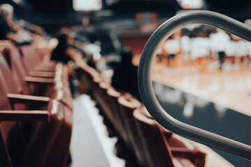 Angled view of empty red stadium seats section with metal safety rails. Blurry players, audience members in background suggest relaxed basketball event in clean venue at college in Dallas, Texas