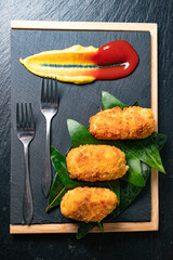 La croqueta se convierte en arte visual: textura, color y gesto culinario en primer plano.Croquetas doradas sobre pizarra negra, hojas verdes, salsas decorativas, tres tenedores metálicos alineados.