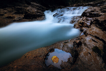 Long exposure of a waterfall in a dark gorge, with a single autumn leaf in a rock puddle.