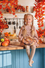 Cute little girl in autumn checkered dress sits on a rustic kitchen counter with fall fruits and Thanksgiving pie, surrounded by bright orange maple leaves decoration